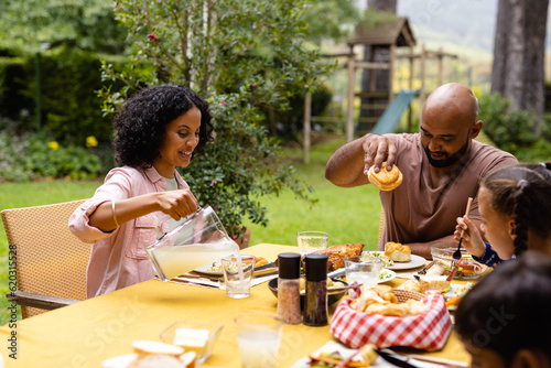 Happy biracial parents, son and daughter sitting at table eating meal in garden, copy space