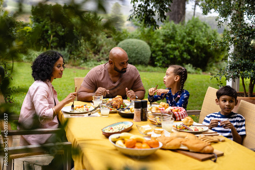 Happy biracial parents  son and daughter sitting at table having meal and talking in garden