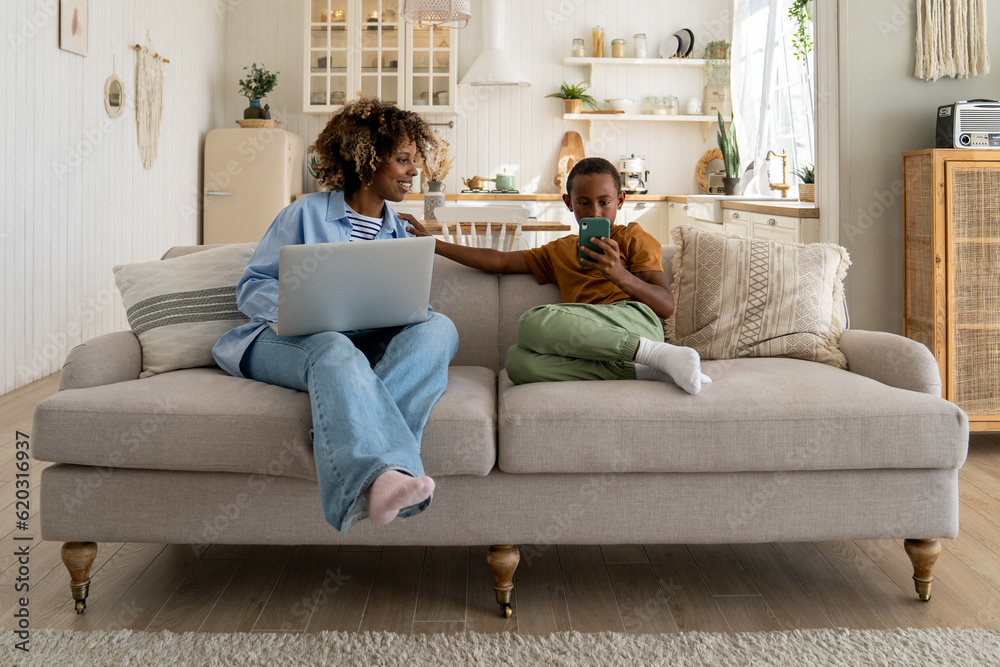 African american mom working on laptop and looking at smartphone in son's hands sitting on couch at home. Teen boy browsing in smartphone, watching videos. Working busy mom, child gadget addiction. African american mom working on laptop and looking at smartphone in son's hands sitting on couch at home. Teen boy browsing in smartphone, watching videos. Working busy mom, child gadget addiction.