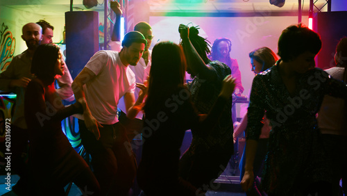 Smiling people dancing at event celebration listening to modern music, partying on dance floor. Diverse adults enjoying dance party with DJ and stage lights, entertainment. Handheld shot.