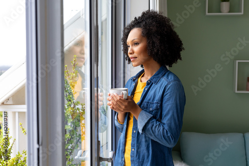 Biracial woman holding cup of coffee and looking out window at home