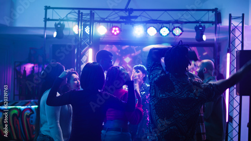 Diverse persons dancing on music, partying with crowd of people and having fun at nightclub with DJ. Group of friends enjoying disco party on dance floor at event. Handheld shot.