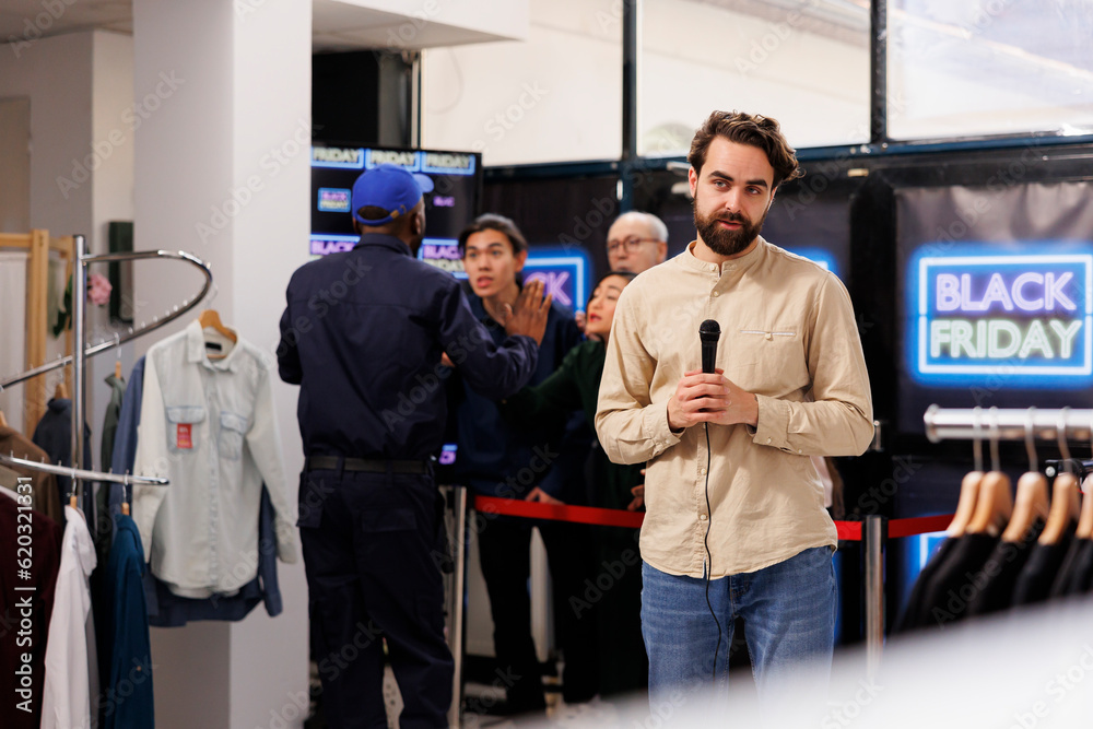 TV reporter working at shopping mall for live shot of people lining up for discounts. Young man journalist holding microphone reporting on retail industry news, broadcasting about Black Friday madness