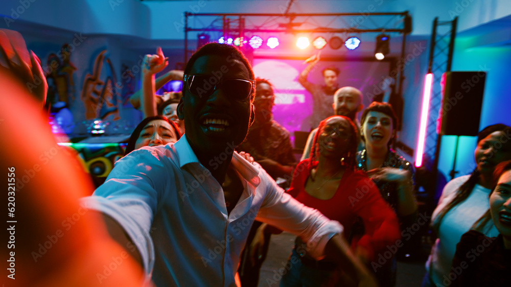Fototapeta premium POV of diverse friends recording video at club, making memories at modern disco party with crowd of people. Adults taking photos and filming vlog at discotheque, dance event. Handheld shot.