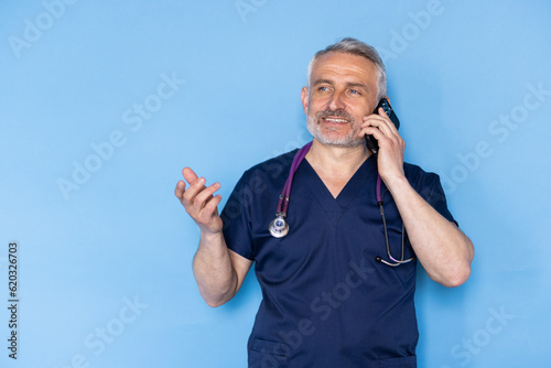 Handsome mature doctor wearing white medical coat using cell phone, calling patient or colleague, blue background