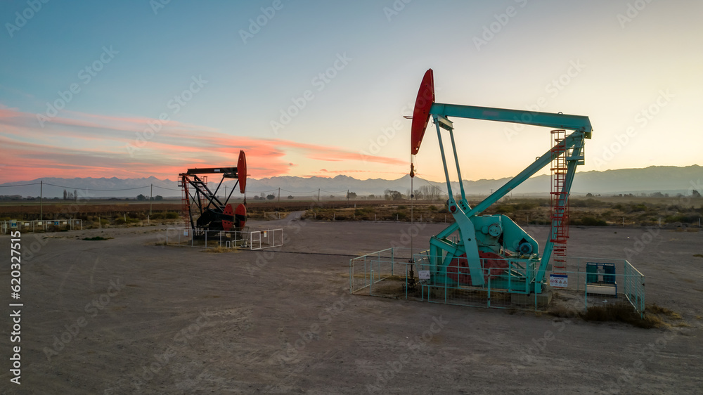 Pumpjack and transmission towers at sunset symbolizing energy transition. A pump jack pumping oil out of a well with silhouettes of electricity pylons and power line against a red sky. Pumpjack and transmission towers at sunset symbolizing energy transition. A pump jack pumping oil out of a well with silhouettes of electricity pylons and power line against a red sky.