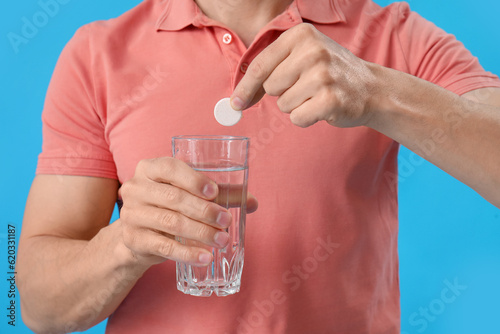 Young man putting soluble tablet into glass of water on blue background, closeup