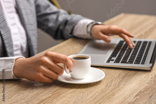 Woman using modern laptop and holding cup of coffee at table