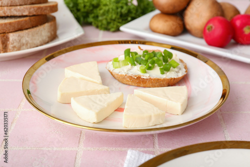Plate with triangles of tasty processed cheese, bread pieces and vegetables on pink tile background, closeup