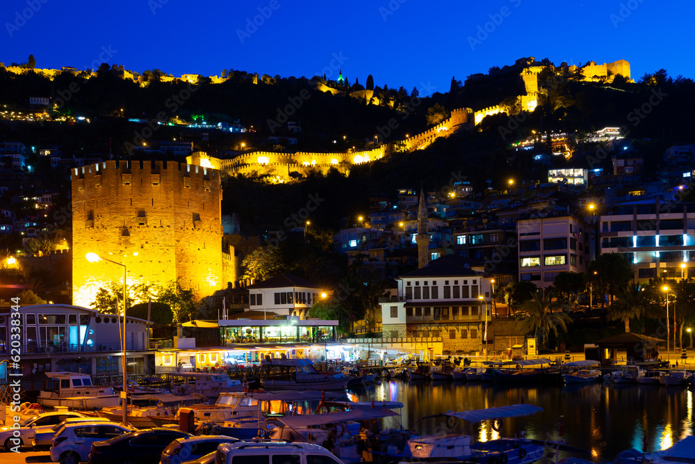 Illuminated pier of Alanya with moored boats in evening. View of Kizil Kule (Red Tower) and Castle of Alanya, Antalya Province, Turkey.