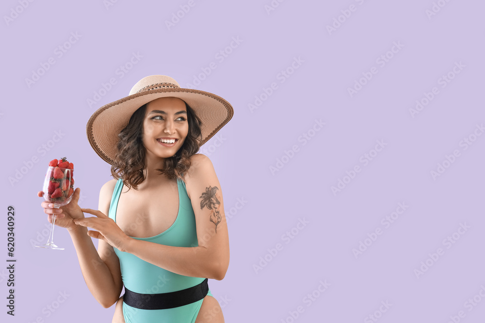 Young woman in swimsuit with glass of strawberries on lilac background Young woman in swimsuit with glass of strawberries on lilac background