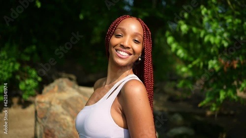 Outdoor portrait of natural Beautiful young African American woman long red braids hair style and perfect white teeth smile, posing in swimsuit at sunny summer day with green foliage beach background