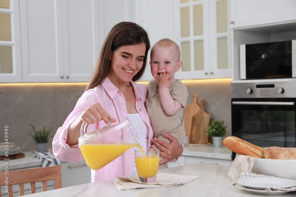 Happy young woman holding her cute little baby while pouring juice into glass in kitchen