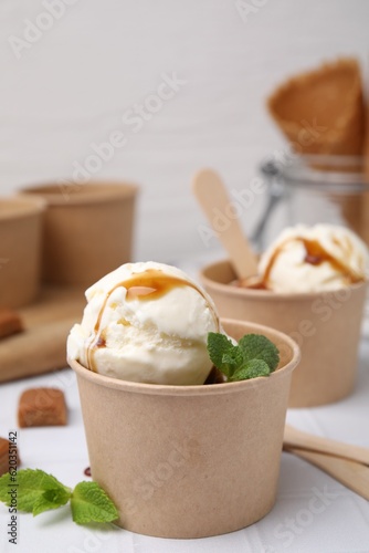Scoops of ice cream with caramel sauce, mint leaves and candies on white tiled table, closeup