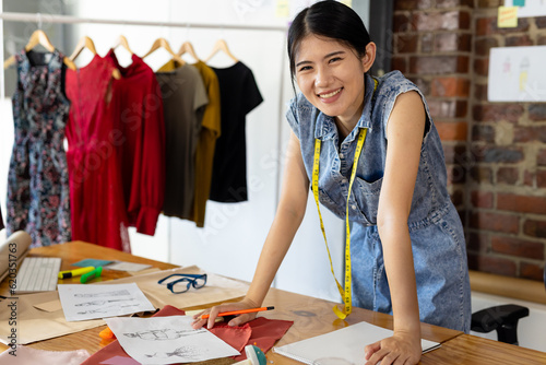 Portrait of asian female fashion designer wearing tape measure leaning on desk smiling at studio