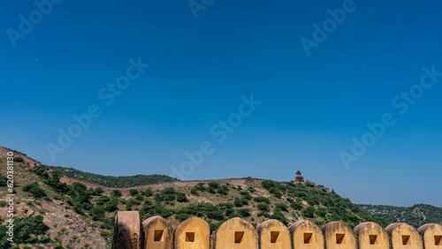 An ancient fortress wall with watchtowers runs along the ridge of the mountain. Clear blue sky. In the foreground is the upper part of the Amber Fort wall. Copy space. Jaipur. India.