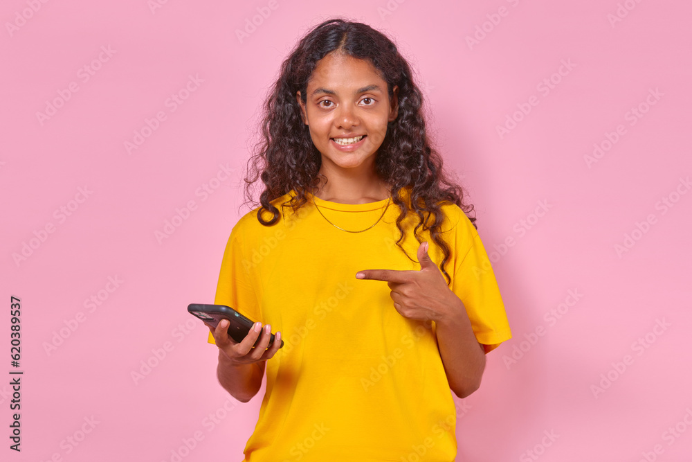 Young positive curly Indian woman teen with smartphone in hand points finger at gadget to recommend you download new online shopping application in stands in pink studio mobile phone. Young positive curly Indian woman teen with smartphone in hand points finger at gadget to recommend you download new online shopping application in stands in pink studio mobile phone.