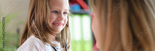 Cheerful small girl using medical stethoscope and looking at doctor with happiness