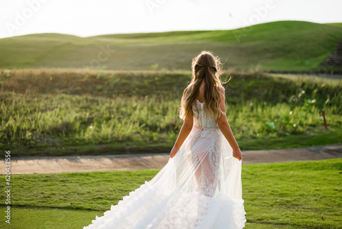 A beautiful, young girl with blond hair runs across the lawn in the light of the setting sun, in a white, stylish wedding dress, rear view.