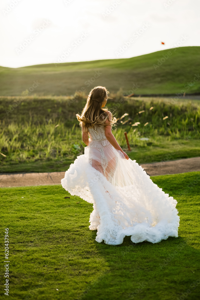 A beautiful, young girl with blond hair runs across the lawn in the light of the setting sun, in a white, stylish wedding dress, rear view. A beautiful, young girl with blond hair runs across the lawn in the light of the setting sun, in a white, stylish wedding dress, rear view.