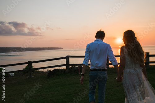  The newlyweds, in beautiful wedding clothes, hold hands and walk along the green lawn, admiring the sunset.