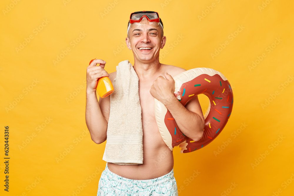 Happy satisfied adult man wearing shorts swimsuit snorkeling goggles holding donut rubber ring and bag isolated on yellow background holding sunscreen for skin care during sunbathing. Happy satisfied adult man wearing shorts swimsuit snorkeling goggles holding donut rubber ring and bag isolated on yellow background holding sunscreen for skin care during sunbathing.