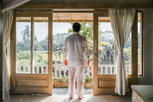 A successful middle-aged man of European appearance, dressed in white, enjoys a gorgeous view of the rice fields from the balcony of his villa.