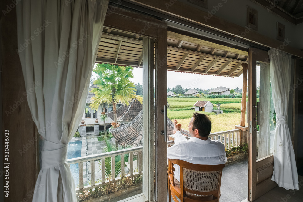 A successful middle-aged man of European appearance works on a laptop, sitting in a villa, drinking wine from a glass, admiring a gorgeous view of the rice fields.