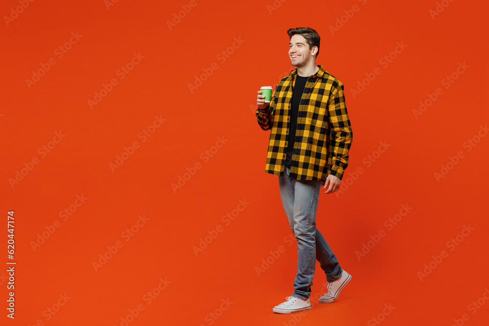 Full body side view young caucasian man he wear yellow checkered shirt black t-shirt hold takeaway delivery craft paper brown cup coffee to go isolated on plain red orange background studio portrait. Full body side view young caucasian man he wear yellow checkered shirt black t-shirt hold takeaway delivery craft paper brown cup coffee to go isolated on plain red orange background studio portrait.