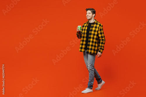 Full body side view young caucasian man he wear yellow checkered shirt black t-shirt hold takeaway delivery craft paper brown cup coffee to go isolated on plain red orange background studio portrait.