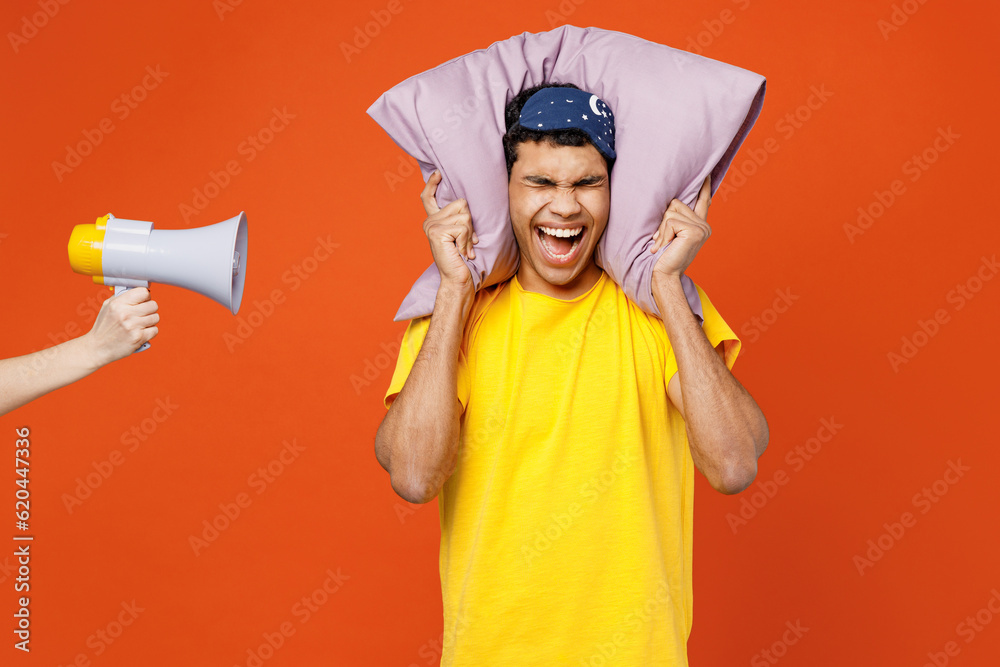 Young man wear pyjamas jam sleep eye mask at home hold pillow behind neck cover ears from neighbours noise, scream in megaphone isolated on plain orange background studio. Bad mood night nap concept. Young man wear pyjamas jam sleep eye mask at home hold pillow behind neck cover ears from neighbours noise, scream in megaphone isolated on plain orange background studio. Bad mood night nap concept.