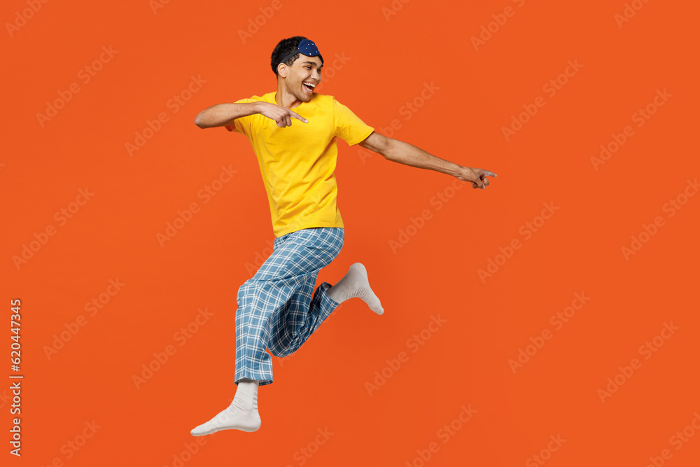 Full body young fun man wear pyjamas jam sleep eye mask rest relax at home jump high point index finger aside on area isolated on plain orange background studio portrait. Good mood night nap concept.
