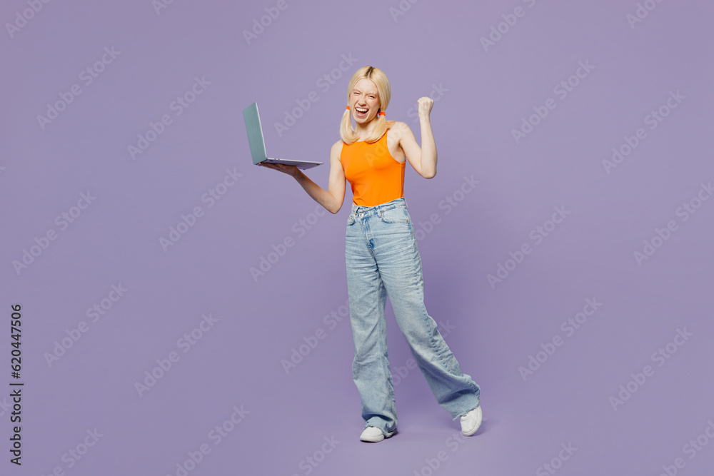Full body young blonde IT woman wear orange tank shirt casual clothes hold use work on laptop pc computer do winner gesture isolated on plain pastel light purple background studio. Lifestyle concept. Full body young blonde IT woman wear orange tank shirt casual clothes hold use work on laptop pc computer do winner gesture isolated on plain pastel light purple background studio. Lifestyle concept.