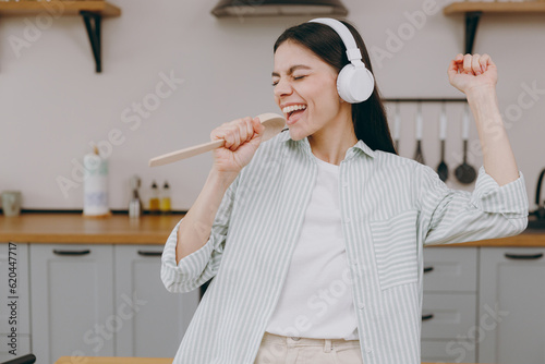 Young housewife latin woman wear casual clothes striped shirt headphones listen to muisc sing song in microphone spoon stand near table in light kitchen at home alone Lifestyle cooking food concept. photo