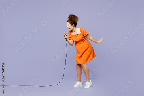 Full body side view young expressive latin woman wear orange blouse casual clothes sing song in microphone at karaoke club isolated on plain pastel light purple background studio. Lifestyle concept photo