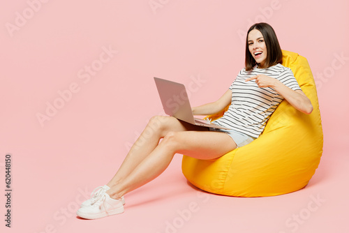 Full body young IT woman in casual clothes t-shirt sit in bag chair hold use work point finger on laptop pc computer isolated on plain pastel light pink background studio portrait. Lifestyle concept