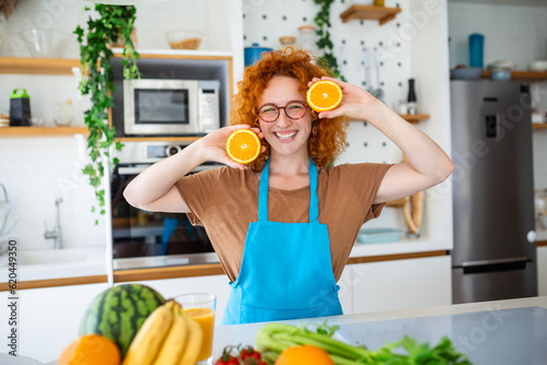 In a modern kitchen, a red-haired woman holds orange slices near her face. With a smile, she embraces the refreshing aroma, radiating vitality and the joy of healthy living in this captivating image.