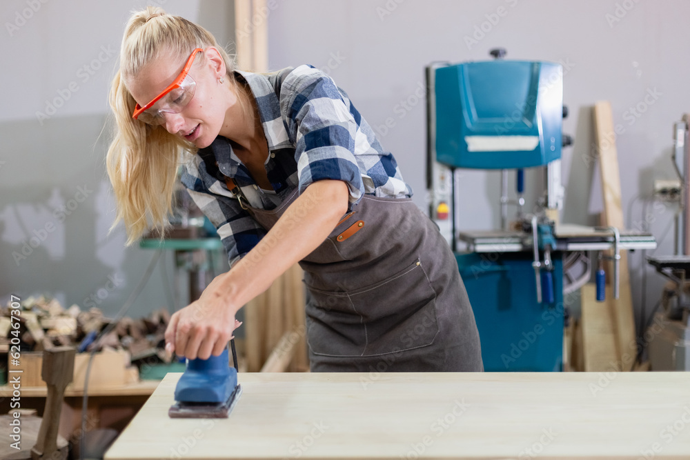 young carpenter caucasian woman using electric wood sander on wood at manufacturing wooden industry. Female craftsman profession in wood factory.