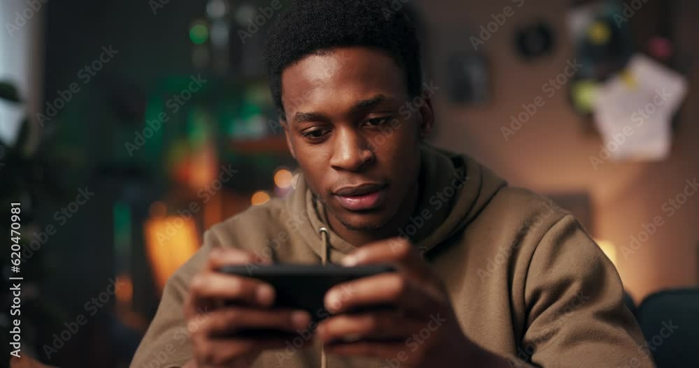 African man sits on a couch in his living room, clutching his cellphone tightly with a look of frustration on his face. He plays a challenging mobile game that causing him stress and disappointment. African man sits on a couch in his living room, clutching his cellphone tightly with a look of frustration on his face. He plays a challenging mobile game that causing him stress and disappointment.