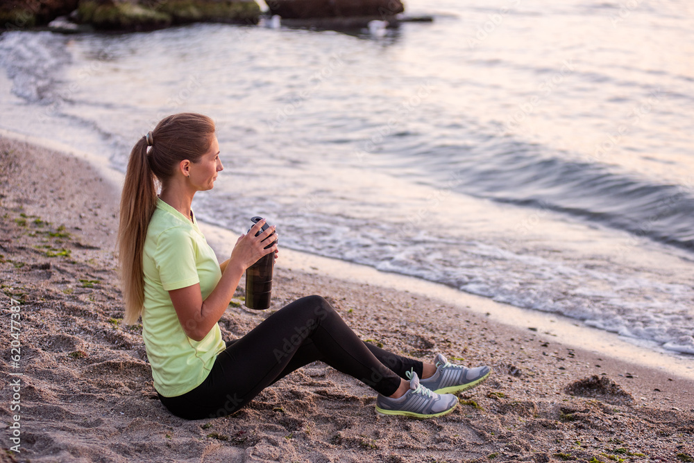 Young woman in sportswear is sitting on the seashore at sunrise. Girl is resting after workout, replenishes her water balance, drinks from a sports bottle, looks at the water. Morning outdoor fitness 