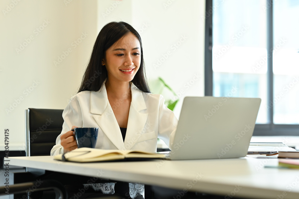 Image of smiling asian woman manager holding cup of coffee and checking business email on laptop computer Image of smiling asian woman manager holding cup of coffee and checking business email on laptop computer