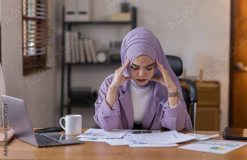 A stressed Asian business asian islamic woman in hijab holding documents is working from home, feeling stressed because of lot of paperwork appearing worried, tired, and overwhelmed