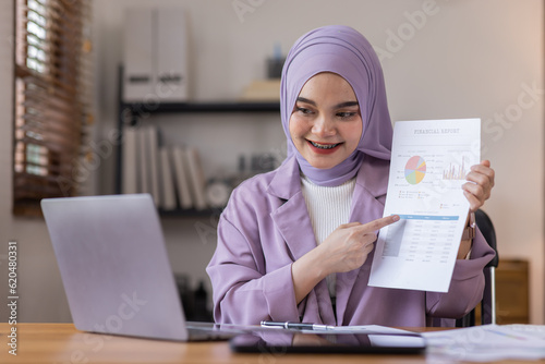 Asian business asian islamic woman in hijab holding financial documents to check calculations on laptop or analyzing report accuracy to show data with graphs to prepare company finances