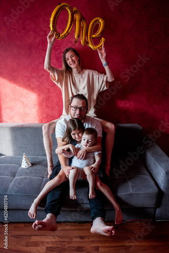 Portrait of happy, diversity family on the birthday of one year old boy. Mother laughs, holds foil balloons ONE in hands, father hugs daughter and son on gray sofa in the sun. Home holiday party.