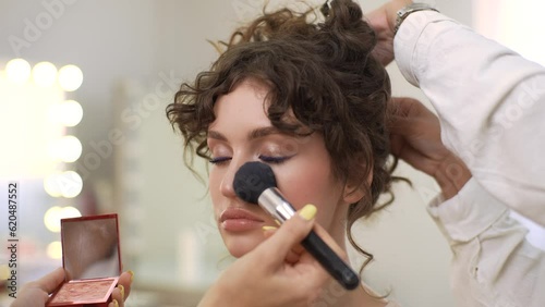 Close-up of unrecognizable wedding makeup artist making fashion make up for beautiful bride in luxury beauty salon. Hairdresser making hairstyle using curling tongs for long hair of female
