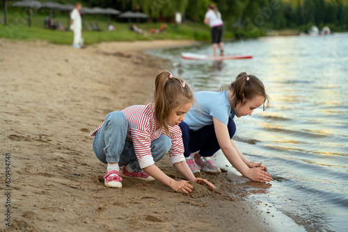 Happy little girls play with sand on river on background of green summer park with people at sunset. Children walk outdoors by water