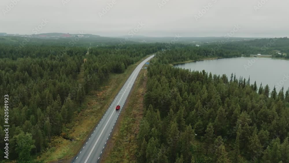 Drone flies directly over a red car driving on a road between a forest and a lake Drone flies directly over a red car driving on a road between a forest and a lake