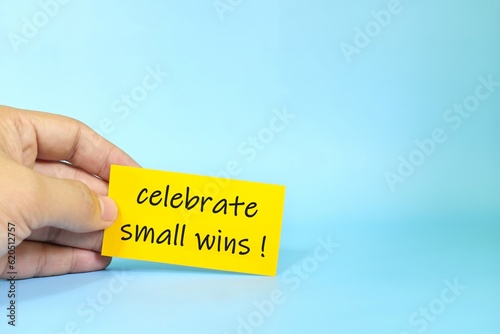 Celebrate small wins reminder concept. Hand holding a bright yellow paper message note.	