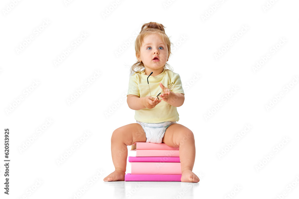 Portrait of curious baby, little girl, child, toddler sitting on books and looking with interest against white studio background Portrait of curious baby, little girl, child, toddler sitting on books and looking with interest against white studio background