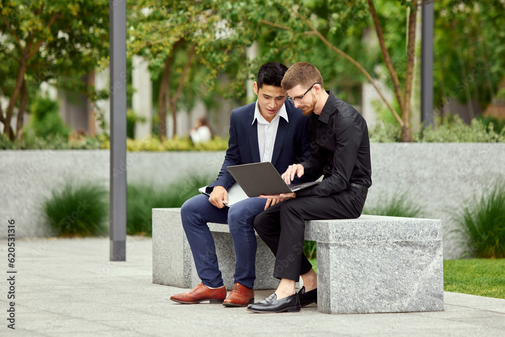 Two businessmen in formal wear sitting outdoors and looking in laptop, working online with projects. Discussion Two businessmen in formal wear sitting outdoors and looking in laptop, working online with projects. Discussion